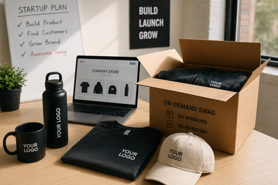 Startup workspace with branded swag including hoodie, cap, mug, and water bottle next to laptop showing company merch store, highlighting on-demand swag with no minimums and zero inventory