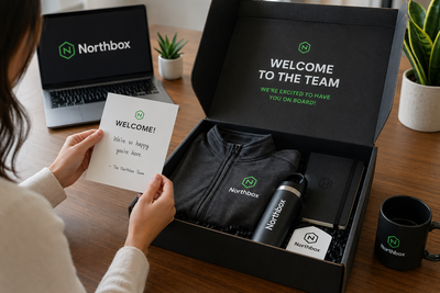 Remote employee opening a new hire welcome kit with branded hoodie, tumbler, notebook, and welcome card on a home office desk, showcasing on-demand onboarding swag for distributed teams